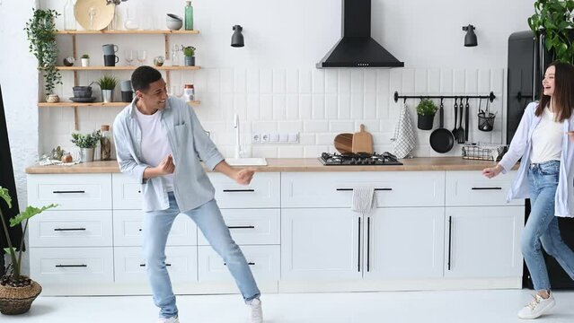 A young modern happy couple in love, newlyweds, stylishly dressed, dance together to their favorite music at home in the kitchen with stylish interior, smile, enjoying to spending time with each other
