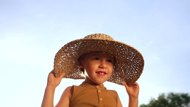 Adorable smiling boy in straw hat on blue sky background. Funny child with kind face looking to camera, enjoying summer outdoors. Childhood, countryside leisure.