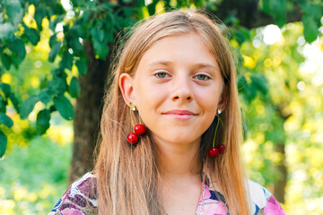 Defocus beautiful blond teen girl with sweet cherry on ear. Beautiful smiling teenage girl closeup, against green of summer park. Child earrings. Summer portrait. Out of focus - Powered by Adobe