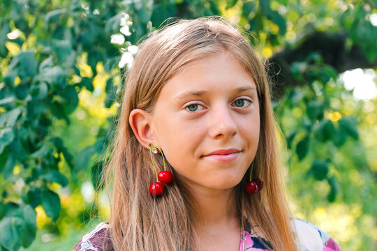 Defocus beautiful blond teen girl with sweet cherry on ear. Beautiful smiling teenage girl closeup, against green of summer park. Child earrings.  Out of focus