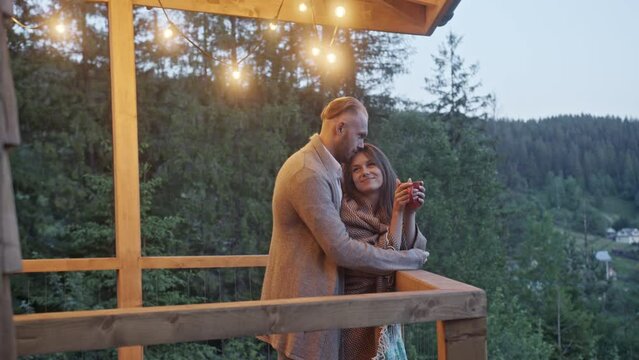 Couple of young married people stands on balcony in evening. Couple of young married people stands on wooden balcony admiring evening nature view. Man gives woman cup of hot tea and hugs against