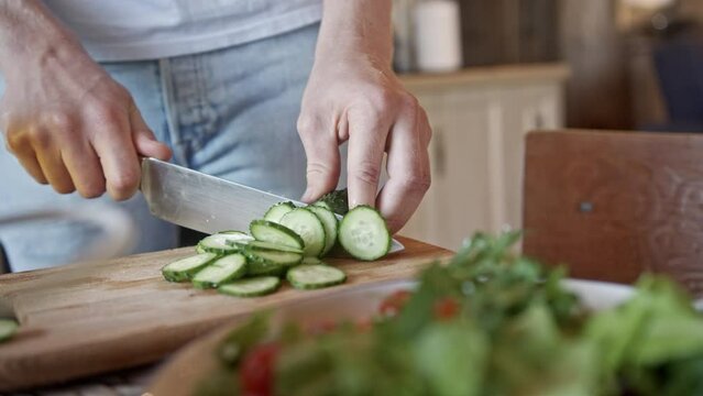 Young male hands cut cucumber using large knife on board. Male hands slice green cucumber using sharp knife on wooden board in kitchen. Preparing salad on kitchen table by cutting vegetables