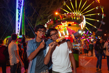 Happy Latino boys eating at the fair with a Ferris wheel behind them. - obrazy, fototapety, plakaty