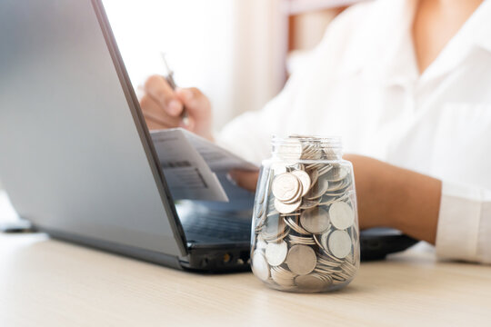 Savings concept coins in jar and woman check expenses list with laptop. Bills payment from savings money plan.
