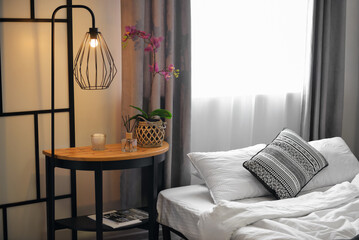 Table with flower, reed diffuser and glowing lamp in stylish bedroom