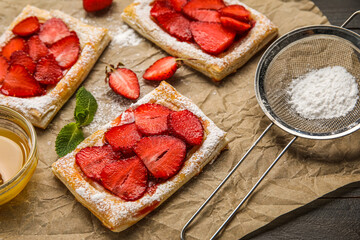 Delicious strawberry puff pastry, bowl with honey and sugar powder on dark wooden table, closeup