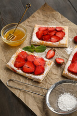 Delicious strawberry puff pastry, bowl with honey and sugar powder on dark wooden table, closeup