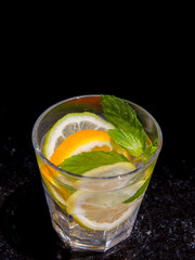 Citrus and mint leaves infused water, cocktail, lemonade in glass. Summer iced cold drink with lemon, lime, orange, mint on black marble background, copy space, top view, vertical