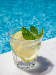 Citrus and mint leaves infused water, cocktail, lemonade in glass near pool. Summer iced cold drink with lemon, lime, ice and mint on blue water white marble background, copy space, side view,vertical
