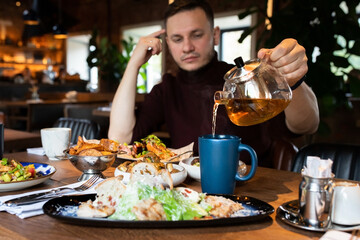 A man in a restaurant pours himself a mug of hot tea from a glass teapot. Front view.