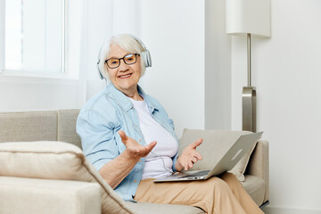 a happy, smiling elderly woman is sitting at home on a cozy sofa with headphones on her head, holding a laptop on her lap while working from home