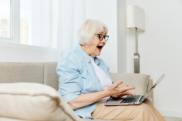 a shocked elderly woman is sitting on a comfortable sofa holding a laptop on her lap and looking very scared at the laptop monitor holding her hand near her face