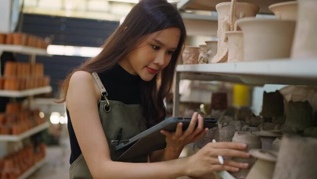 Asia female ceramist with apron hand arrange her clay sculp product on the shelf waiting for bake in oven,asian woman small business ceramic workshop owner smiling proud to present her new collection 