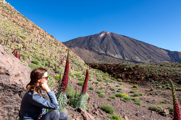 Tourist woman sitting in field of red flowers Tajinaste. Scenic view on volcano Pico del Teide, Mount El Teide National Park, Tenerife, Canary Islands, Spain, Europe. Hiking trail on sunny summer day