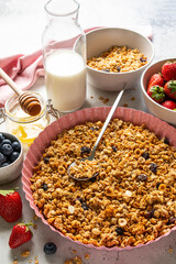 Homemade granola with raisin, seeds, hazelnut and peanut on baking sheet, milk or yogurt bottle, honey and blueberries, strawberries on grey background. Healthy breakfast ingredients. Selective focus.