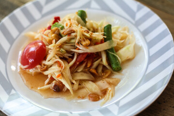 Thai papaya salad on a plate, papaya salad with poop, lemon, pepper, red tomato in white bowl on wooden table