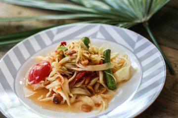 Thai papaya salad on a plate, papaya salad with poop, lemon, pepper, red tomato in white bowl on wooden table