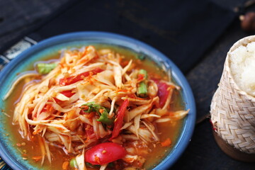 Spicy papaya salad served in a blue dish with sticky rice in a bucket and long beans, dried chilli and vegetables.