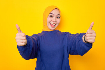 Smiling young Asian Muslim woman dressed in casual sweater showing thumbs up gesture with hand, recommending something good isolated over yellow background