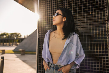 Image of beautiful fair-skinned woman in sunglasses staying on the street . Brunette chinese lady with keeps hands in jeans pockets. City life concept 