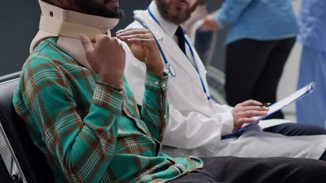 African american patient with medical neck collar talking to physician in hospital reception waiting area. Man with injury after accident wearing cervical brace foam to cure back trauma.