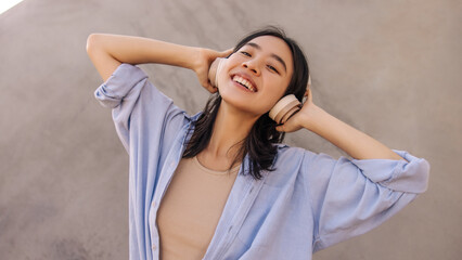 Portrait of happy cute asian woman in headphones look away . Smiling brunette keep hands on head staying on the street. Leisure of life, technology 