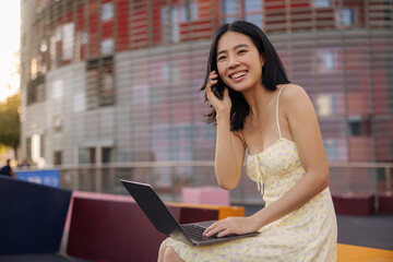 Charming pretty young woman smiling and talking on phone . Fair-skinned chinese sitting with laptop in the city looking away. Lifestyle, technology concept 