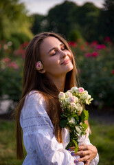 Naklejka premium portrait of a beautiful brunette smiling woman in white embroidered shirt and jeans shorts with bouquet of flowers in summer rose park