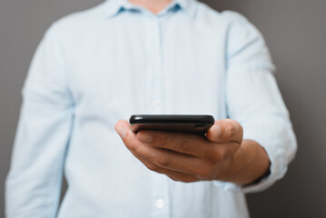 Unrecognizable man in blue shirt holding smartphone while standing against gray background indoors, close-up. Front view businessman showing mobile phone. Selective focus on device