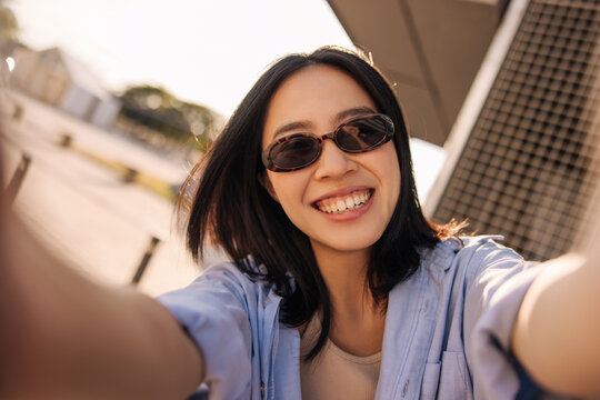 Picture of beautiful young chinese woman in sunglasses in the city . Dark short hair lady look with smile at camera and doing selfie. Concept of use technology 