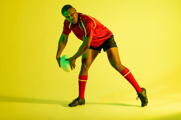 African american male rugby player with rugby ball over yellow lighting