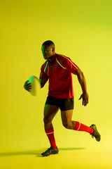 African american male rugby player running with rugby ball over yellow lighting