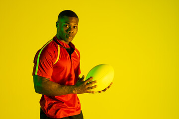 Portrait of african american male rugby player with rugby ball over yellow lighting