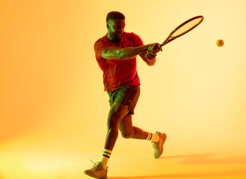 Image of african american male tennis player hitting ball in yellow lighting