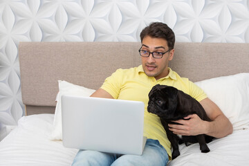 Surprised happy man sitting on the bed and hugging his lovely pet black pug breed and working on laptop. Friendship concept