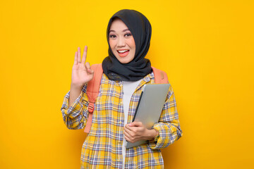 Smiling young Asian Muslim woman student in plaid shirt and backpack hold laptop pc computer, doing approval ok gesture with finger isolated on yellow background