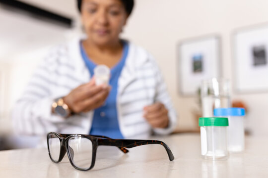 Biracial mature woman with eyeglasses and medicine bottles on table at home