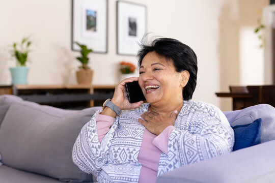 Biracial mature woman with short hair laughing while talking over smartphone and relaxing on sofa