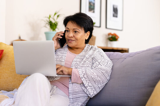 Biracial mature woman with short hair talking over cellphone and using laptop while relaxing on sofa