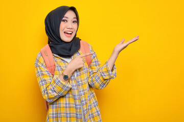 Cheerful young Asian Muslim woman student in casual clothes and backpack, showing copy space on palm isolated on yellow background.Education school university college concept