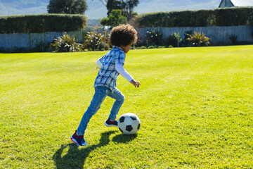 Side view of biracial boy with afro hair running and kicking soccer ball on grassy field in yard - Powered by Adobe
