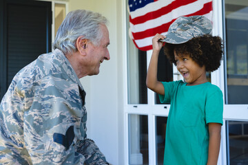 Caucasian military grandfather looking at cheerful biracial boy wearing camouflage cap outside house