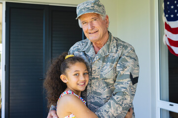 Portrait of smiling multiracial military grandfather in camouflage clothing embracing granddaughter