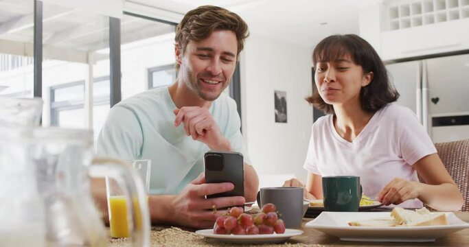 Video of happy diverse couple using smartphone and drinking coffee together in kitchen