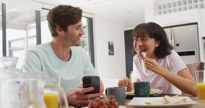 Video of happy diverse couple using smartphone and drinking coffee together in kitchen