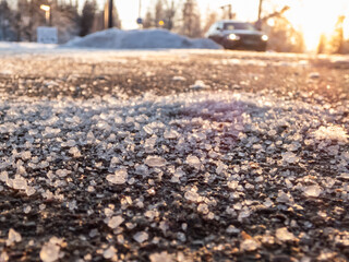 Salt grains on icy sidewalk surface in the winter. Applying salt to keep roads clear and people safe in winter weather from ice or snow. Macro view of salt grains with winter scenery in bacground