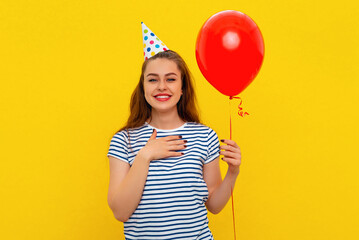 Beauty brunette girl in a birthday cap hold arm pressed to heart and sighing from admiration and delight, feel thankful for the congratulations, holds red inflated helium balloon. Holiday concept
