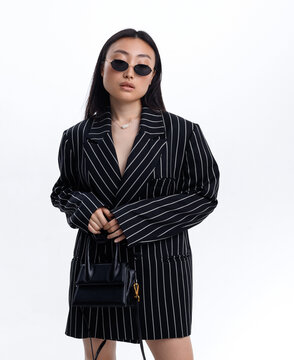 Beautiful Asian girl in black striped jacket wearing sunglasses posing against white wall in photo studio. Fashion shooting