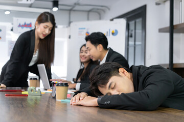 An Asian businessman sleeps in the back of his office as his colleague is working diligently. The concept of taking advantage of colleagues exhaustion at work overwork