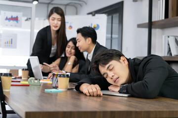 An Asian businessman sleeps in the back of his office as his colleague is working diligently. The...